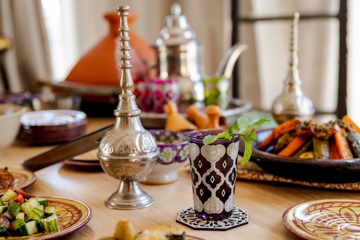 A Moroccan table set for guests: a clay tagine, a silver teapot and rosewater sprinklers, painted plates and salads.