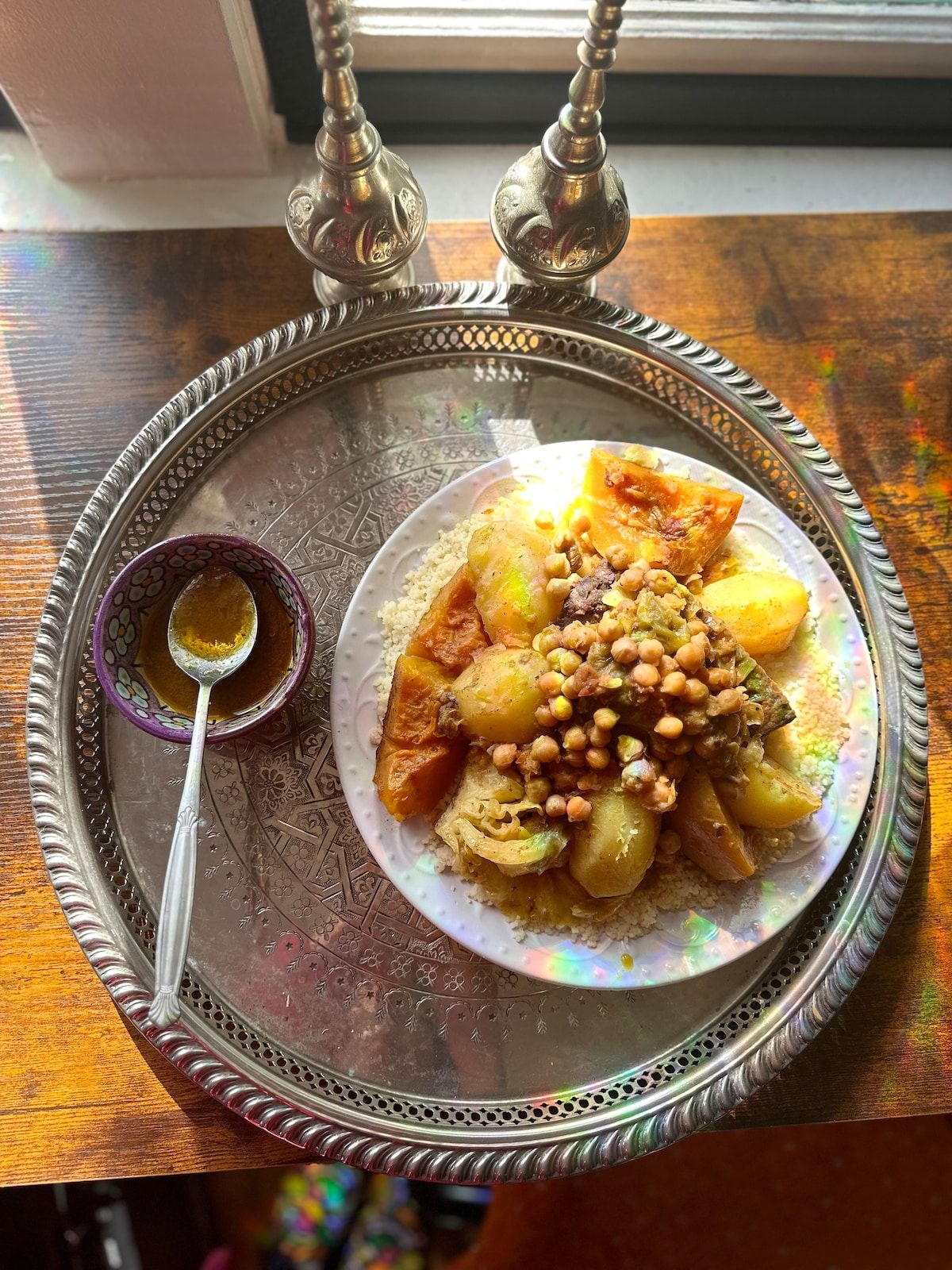 Moroccan couscous topped with chickpeas, pumpkin, potatoes and cabbage, with a small bowl of broth, on a silver tray.