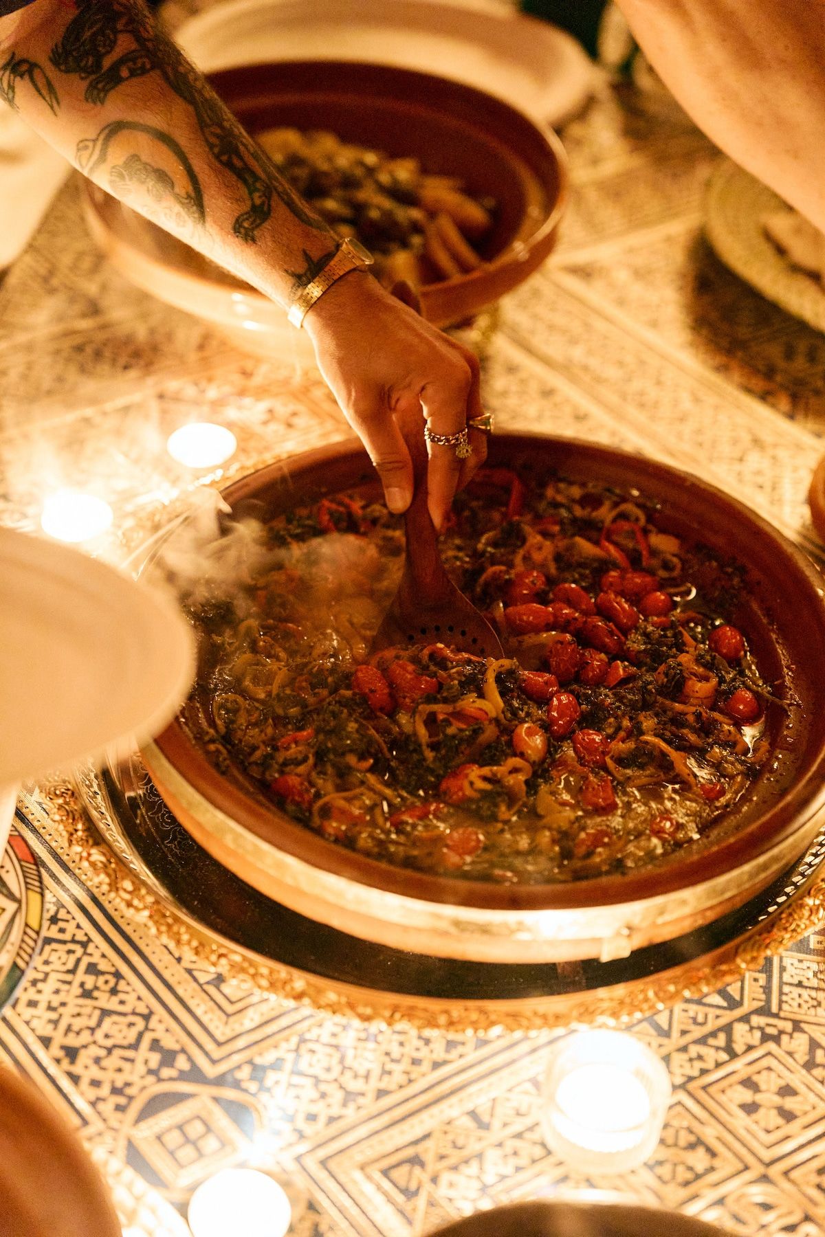 A hand with gold rings stirring a steaming tagine of peppers, cherry tomatoes and herbs with a wooden spoon, among tea lights on a patterned tablecloth.