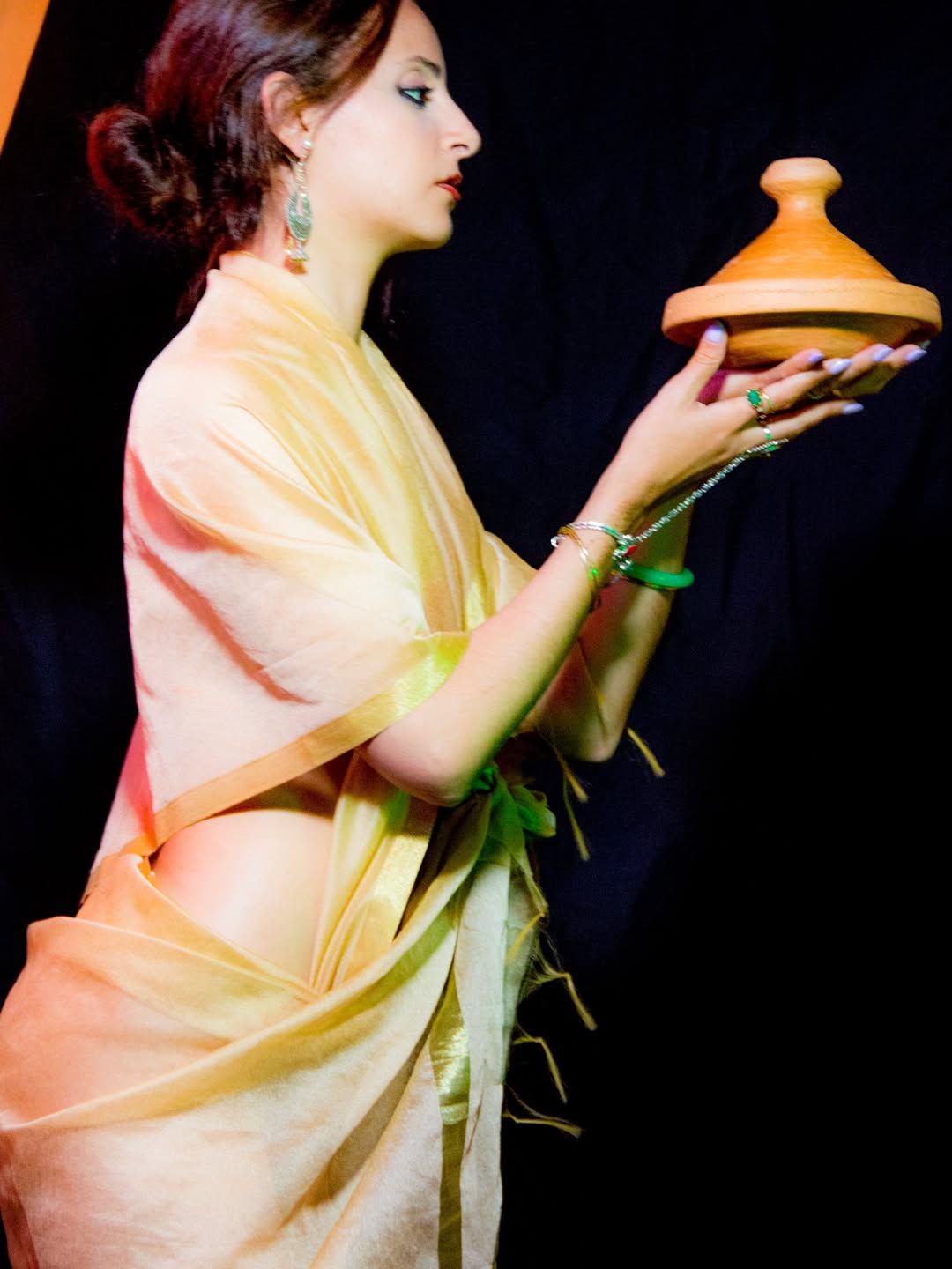 Chef Basmah in a golden silk wrap, in profile, holding up a clay tagine.