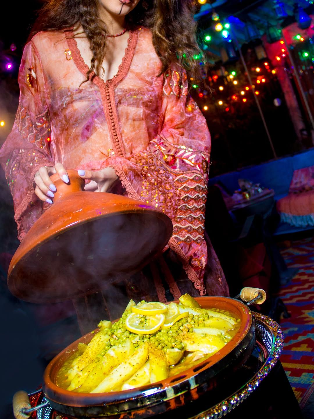 Chef Basmah in a pink kaftan lifting the lid of a fish tagine with lemon slices.