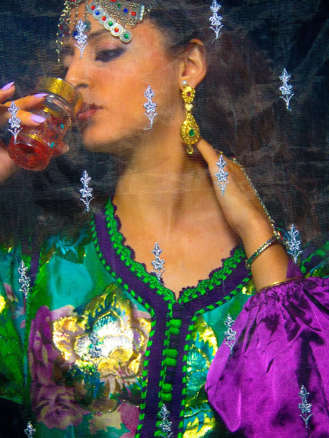 Chef Basmah in a green kaftan and silver headpiece sipping mint tea behind an embroidered veil.