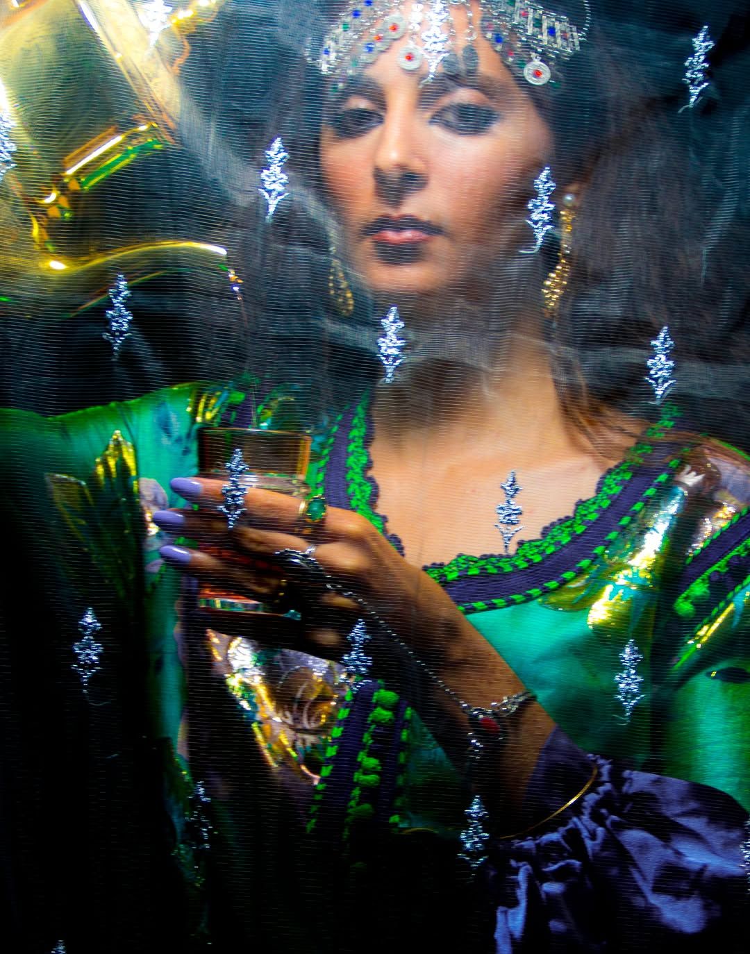 Chef Basmah in a green kaftan and silver headpiece holding a glass of tea behind a sheer veil.