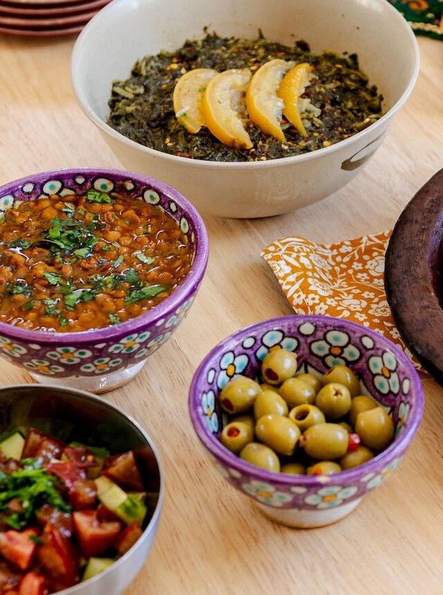 Vegan Moroccan sides in painted bowls: lemony spinach bekkoula, lentil stew, green olives and a tomato-cucumber salad.